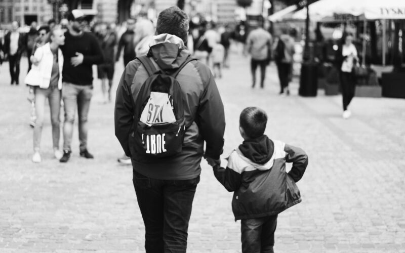 Father and son holding hands while walking through a busy city street in grayscale.