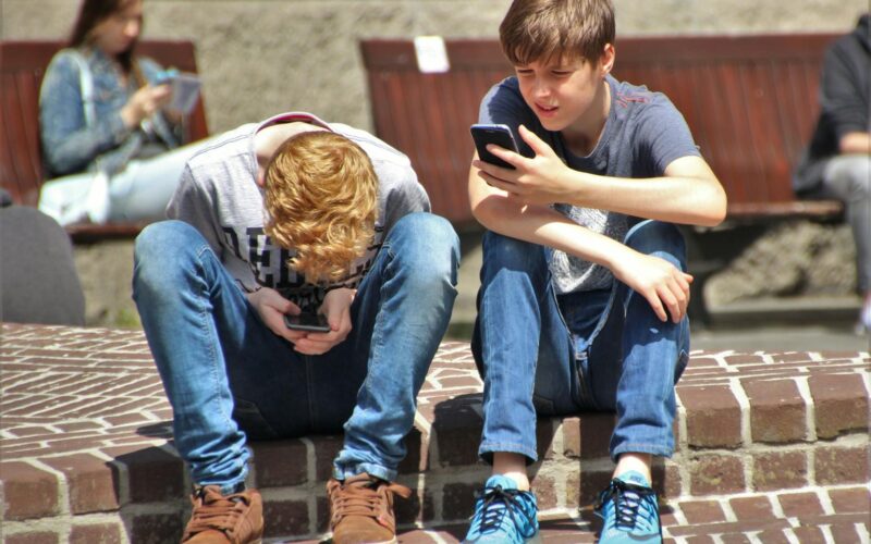 Two teenagers seated outdoors, focused on their smartphones, embracing modern technology.