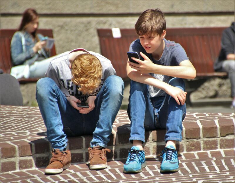 Two teenagers seated outdoors, focused on their smartphones, embracing modern technology.