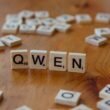 Wooden Scrabble tiles form the word 'QWEN' on a wooden surface, with scattered tiles in the background.