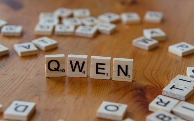 Wooden Scrabble tiles form the word 'QWEN' on a wooden surface, with scattered tiles in the background.