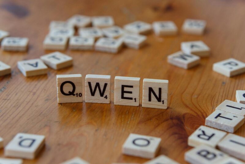 Wooden Scrabble tiles form the word 'QWEN' on a wooden surface, with scattered tiles in the background.