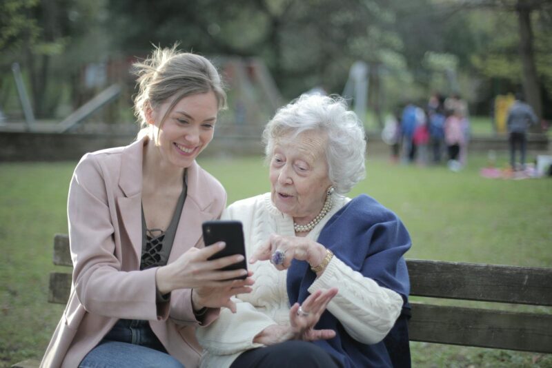 Femeie în vârstă împreună cu nepoata sa, pe o bancă în parc, în timp ce îi arată ceva pe telefon