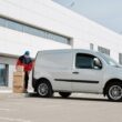 A delivery driver wearing a face mask unloads packages from a van outside a modern building.