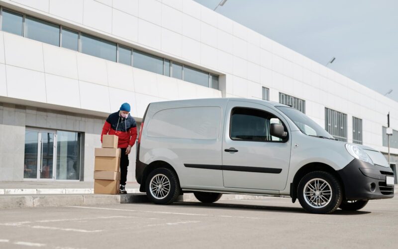 A delivery driver wearing a face mask unloads packages from a van outside a modern building.