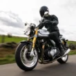 motocicleta clasica cu rider imbracat complet in negru, suprinsa in viteza cu un camp verde in fundal