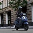 Scuter albastru surprins pe strazile unui oras cu o cladire de culoare gri in partea dreapta