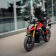 motocicleta rosie pe strada in fata unei cladiri din sticla cu un rider imbracat in negru