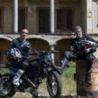 doi motociclisti langa o motoreta neagra cu o cladire parasita in spate