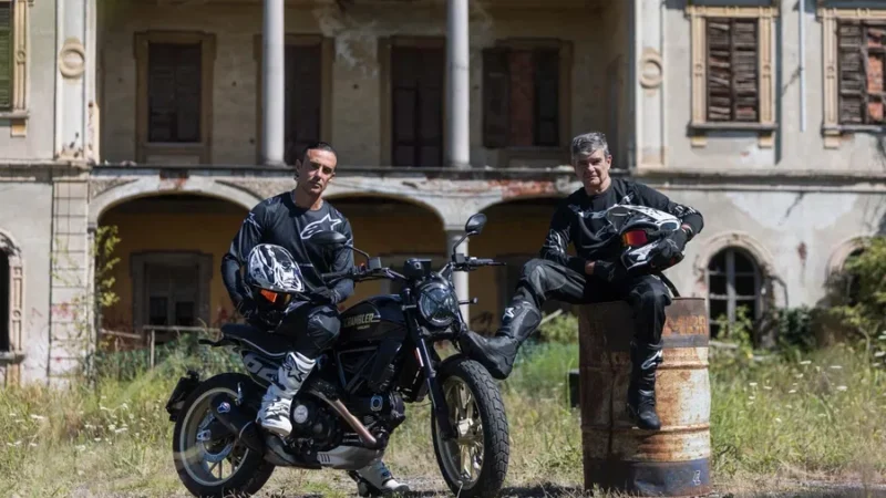 doi motociclisti langa o motoreta neagra cu o cladire parasita in spate