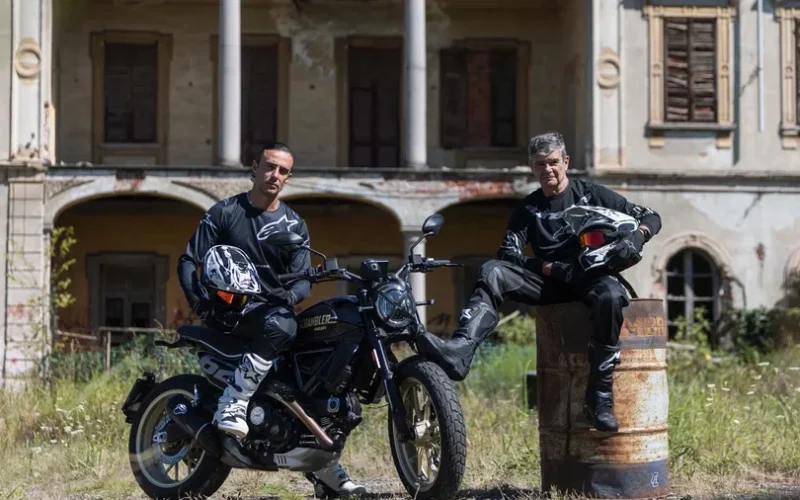 doi motociclisti langa o motoreta neagra cu o cladire parasita in spate