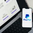 Smartphone cu logo-ul Paypal langa un laptop deschis pe un site de cumparaturi