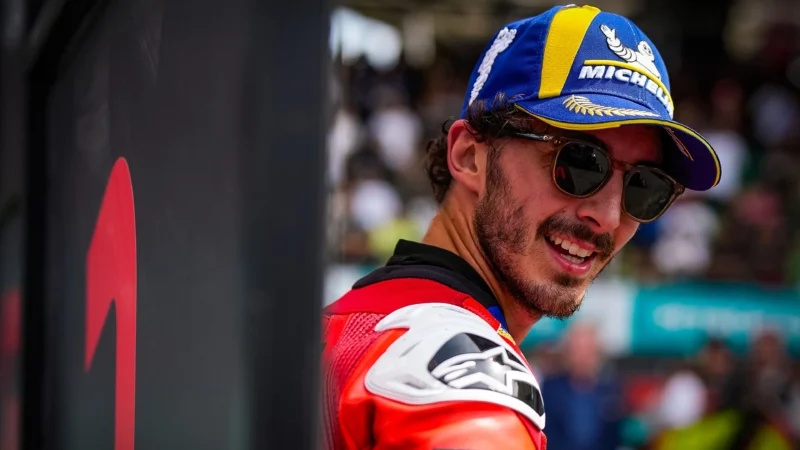 pilot motogp cu o sapca albastra pe cap si ochelari in combinezon rosu cu multime in fundal