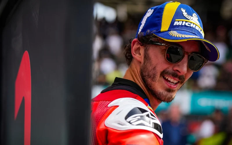 pilot motogp cu o sapca albastra pe cap si ochelari in combinezon rosu cu multime in fundal