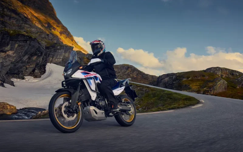 motocicleta honda andventure de culoare alb albastra pe un drum virajat cu un munte in fundal