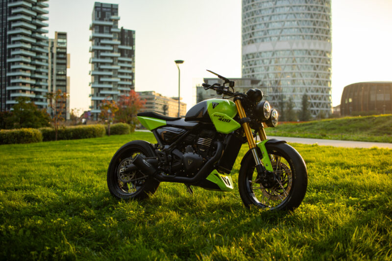 motocicleta verde de tipul naked pe un camp cu cladiri in spate